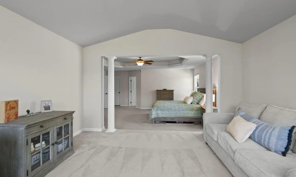 This is an image from Goode Design Painting & Wallcovering Inc., located in Virginia. The photo shows a spacious bedroom with soft neutral walls, white columns, and a sitting area with light-colored furniture.