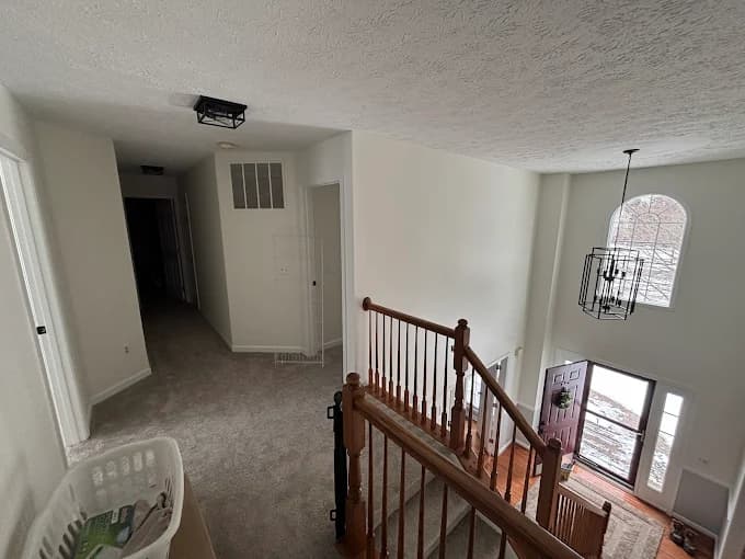 This is an image from Bates Brothers Painting LLC, located in New York. A freshly painted upper hallway and two-story foyer with clean white walls, wooden banister railing, and a modern chandelier by the front entrance.