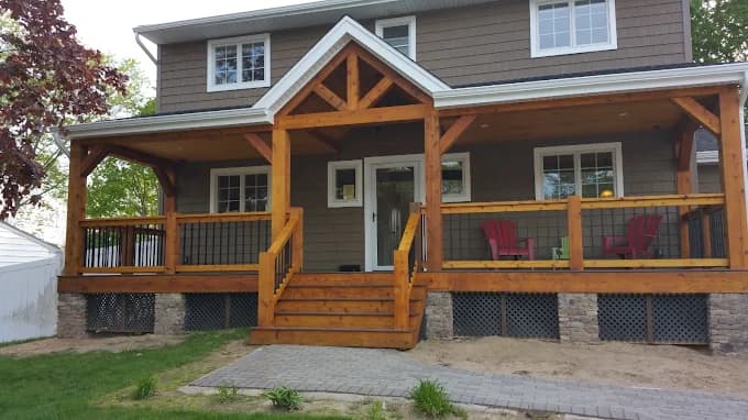 This is an image from Todd's Painting, located in New York. The photo shows a large wood-stained covered porch with timber framing, railing details, and stonework along the foundation.