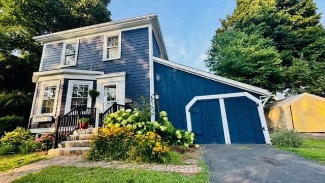 This is an image from CertaPro Painters of North Shore and Cape Ann, located in Massachusetts. It shows a two-story house painted in deep blue with white trim, complemented by a neatly landscaped yard and blooming flowers.