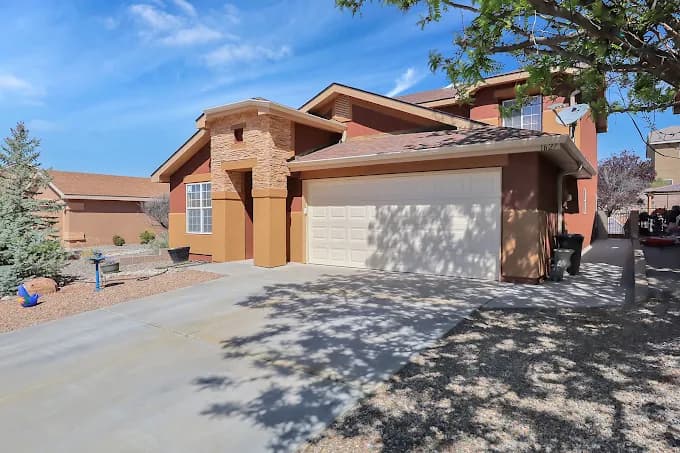 This is an image from Mike’s Quality Painting, located in New Mexico. The photo shows a freshly painted tan stucco two-story home with white trim, a two-car garage, and a clean desert landscape under a bright blue sky.