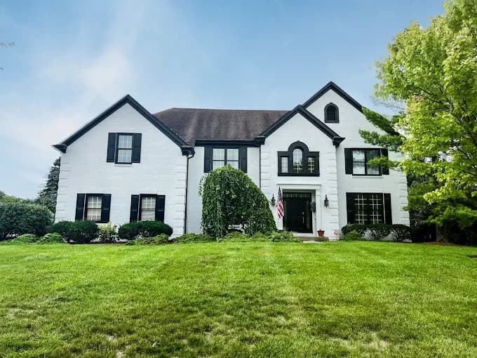This is an image from Spray-Net Lexington, located in Kentucky. The image shows a newly painted white two-story home exterior with black shutters, manicured landscaping, and a bright, uniform finish.