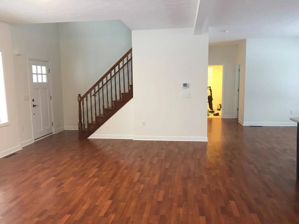 This is an image from Roman Paint Pros company, located in Penn Hills, PA. The image is a freshly painted living room with white walls, wood staircase, and polished hardwood floors in a modern home by Roman Paint Pros.