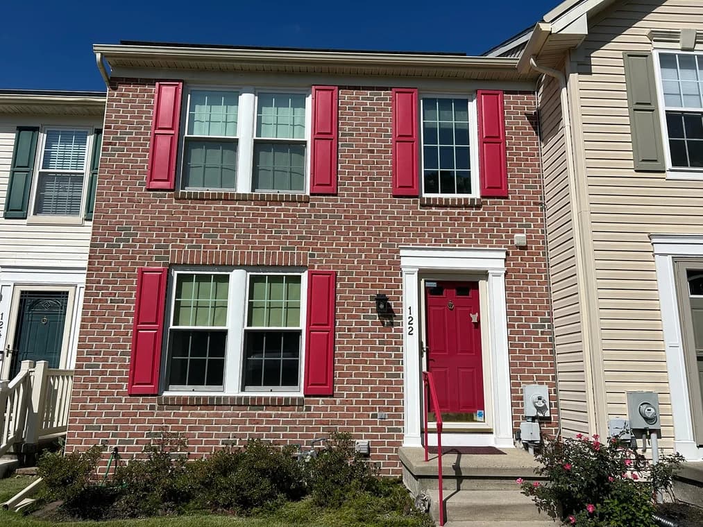 This is an image from Clean Lines Painting & Powerwashing of New Jersey, the image is a brick townhouse with red shutters and a matching red front door, freshly painted by Clean Lines Painting + Powerwashing, giving the home a bold and stylish look.