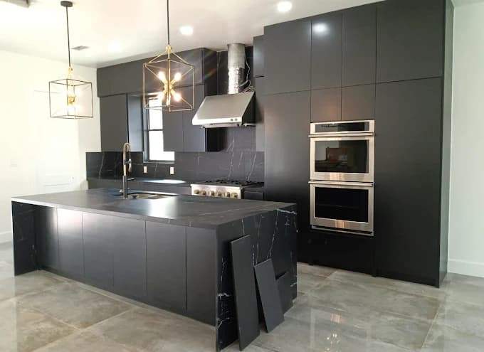 This is an image from Blue Painting, located in Texas. The image shows a modern kitchen with black cabinetry, a large black island with waterfall edges, stainless steel appliances, and two geometric pendant lights above polished tile flooring.
