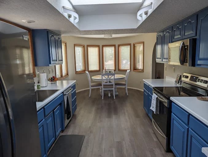 This is an image from Mike’s Quality Painting, located in New Mexico. The photo shows a modern kitchen with freshly painted blue cabinets, white countertops, stainless steel appliances, and a bright dining area with wood-trimmed windows and natural light from a skylight.