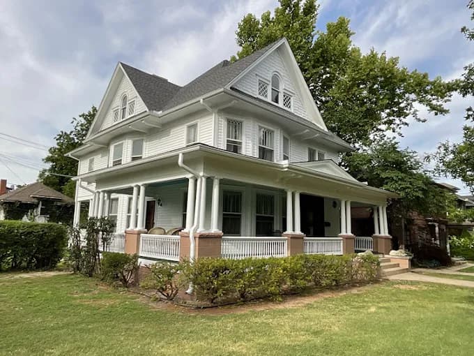 This is an image from CertaPro Painters of OKC, located in Oklahoma. The photo shows a large historic-style two-story home painted white with gray roofing, a wraparound porch, and detailed trim work.