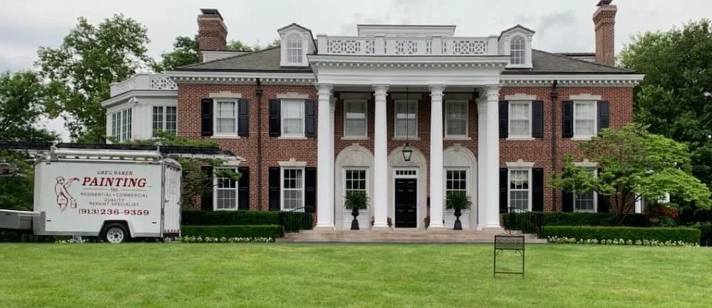 This is an image from Greg Baker Painting. The image is exterior painting in Kansas City KS showing a large brick colonial-style home with tall white columns and black shutters.