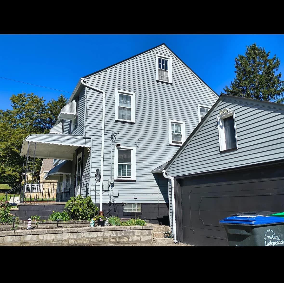 This is an image from 216 Painting, located in Ohio. The image shows a freshly painted gray home exterior with white trim, a black garage door, and landscaped front yard under a clear blue sky