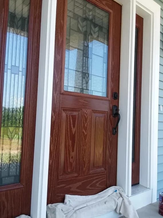 This is an image from Staib & Son Painting & Decorating LLC company, located in Jackson, MI The image is abeautifully refinished front entry door with stained wood grain finish, decorative glass panels, white trim, and freshly painted siding.