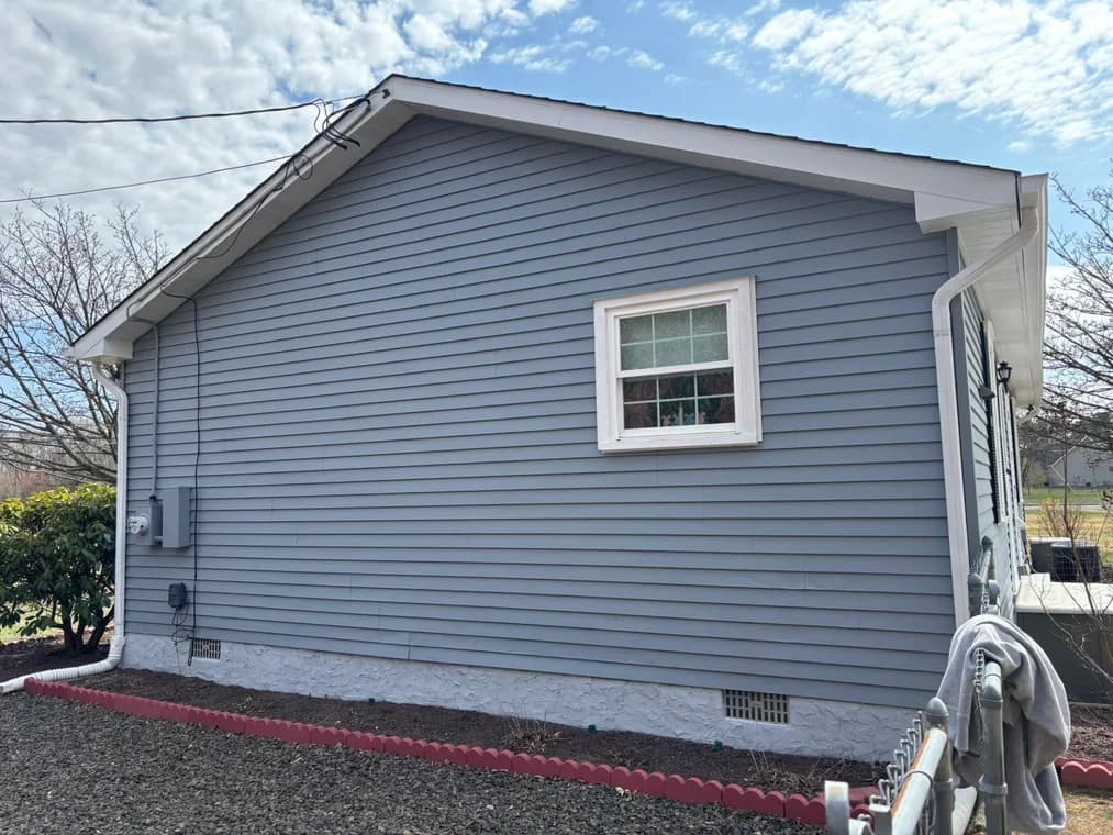 A single-story house with light blue-gray siding and white trim freshly painted by WallPro Painting & Design, featuring a neat exterior with landscaped edging.