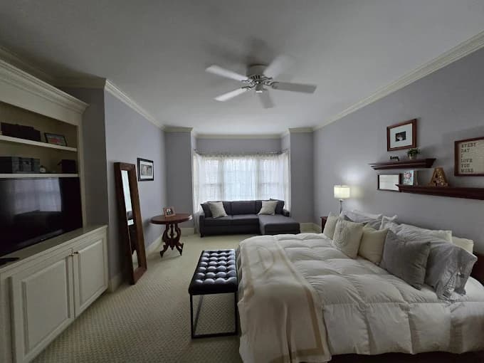 Interior painting project by Johnson Painting Co. & More in Kernersville, North Carolina. The image shows a gray bedroom with crown molding, a ceiling fan, shelves, and a made bed.
