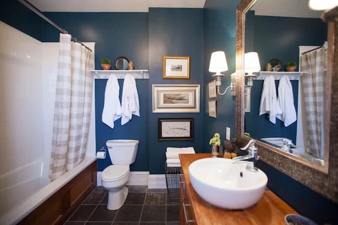 This is an image from Kennedy Painting Interior & Exterior, located in Indiana. The image shows a freshly painted bathroom interior with deep blue walls, white trim, a wood vanity, and clean edges demonstrating detailed interior painting services.