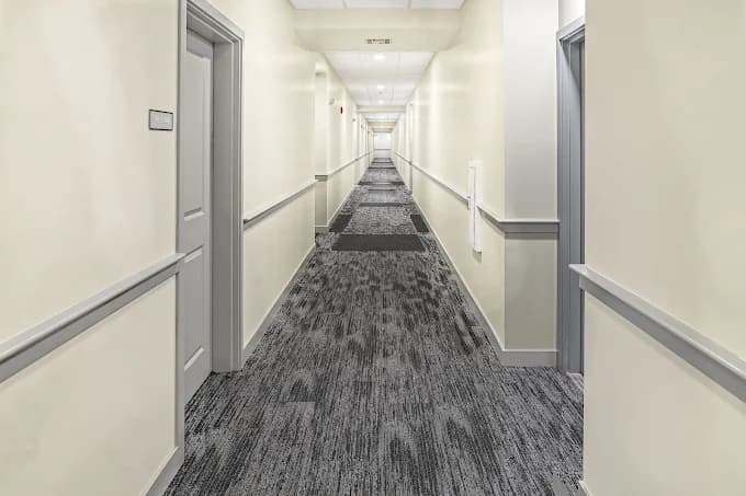 This is an image from Gonnella Painting & Restoration, located in Massachusetts. It shows a long, freshly painted apartment hallway with cream-colored walls, gray trim, and patterned carpeting.