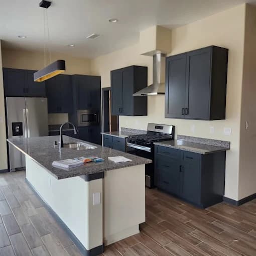 This is an image from Mike’s Pro Painting, LLC, located in New Mexico. The photo shows a newly painted modern kitchen with dark gray shaker cabinets, stainless steel appliances, granite countertops, and wood-look tile flooring.