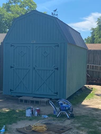 A small backyard shed painted in a muted blue shade, with tools and paint cans visible nearby, completed by Groovy Hues Painting of Tulsa.
