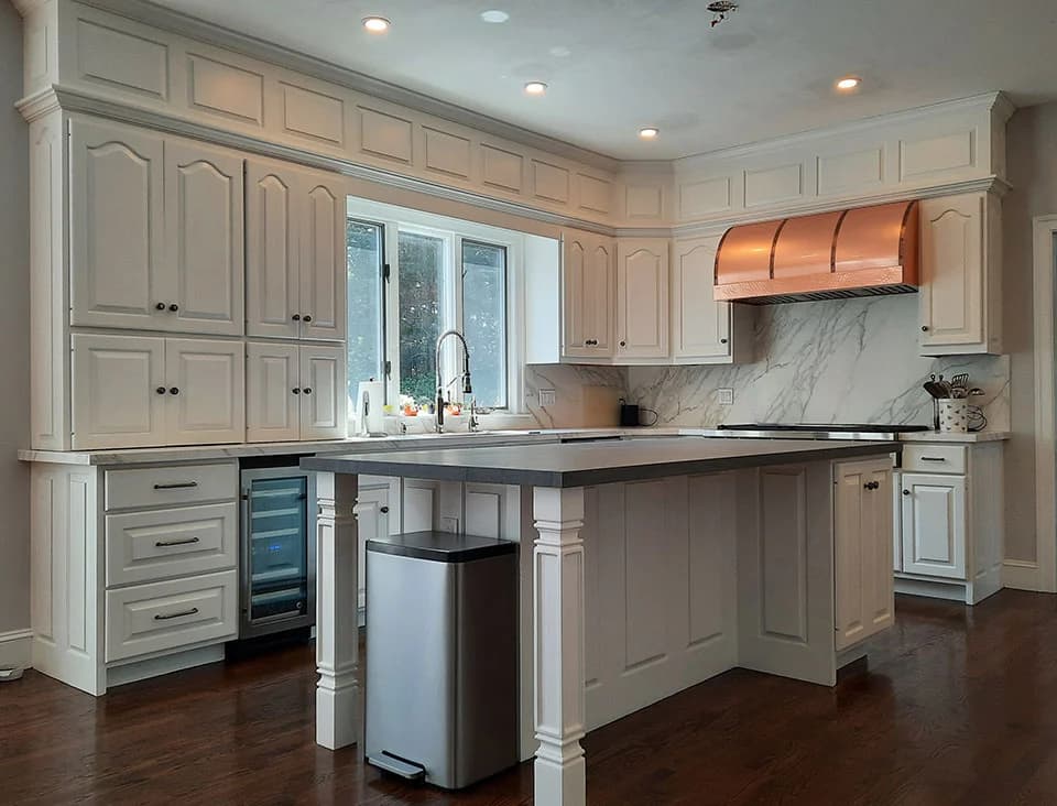 This is an image from Linda Loo Designs Cabinet Refinishing & Refacing, displaying a large kitchen with white cabinetry, a dark countertop, and a copper range hood above the stove. The space looks bright with natural light from a window.