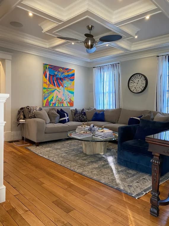 This is an image from CRD Painting Inc, located in New York. The photo displays a furnished living room with detailed coffered ceilings, light gray walls, warm wood flooring, and a large sectional sofa.