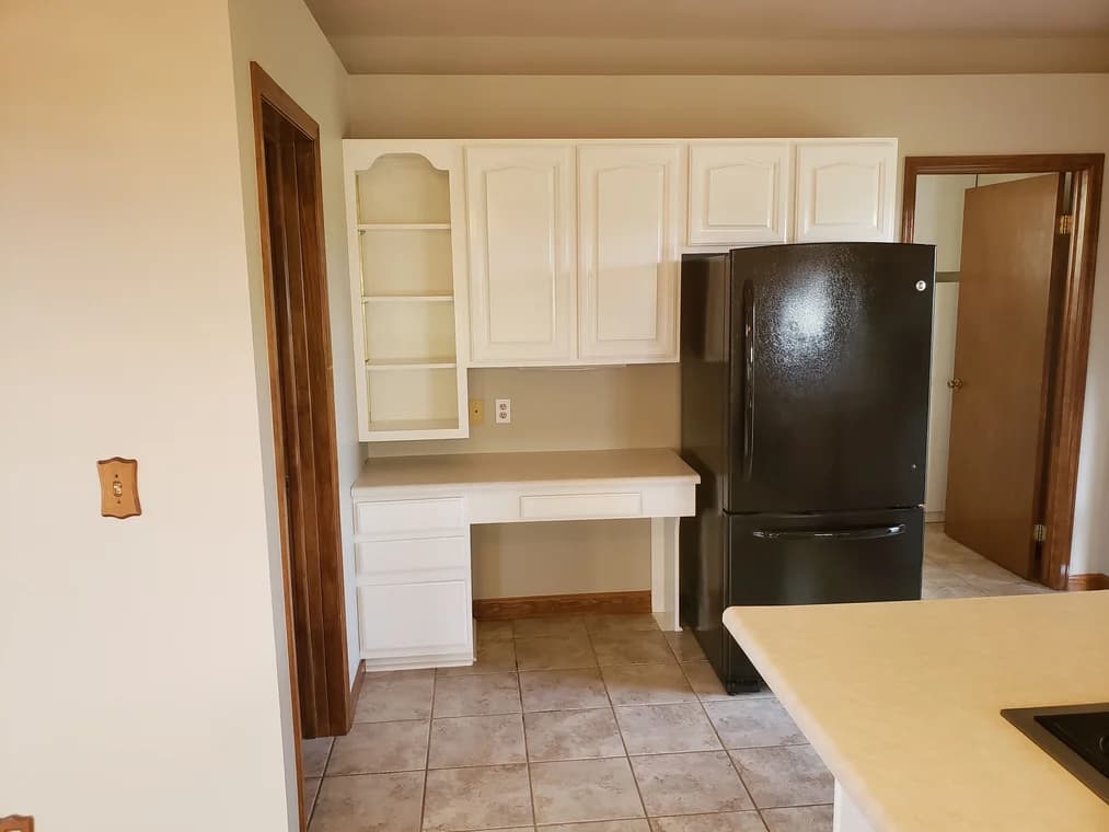 This is an image from Brush Brothers Painting LLP company, located inGolden, CO The image is a freshly painted white kitchen cabinets with black refrigerator and built-in desk area on tile floor by Brush Brothers Painting LLP.