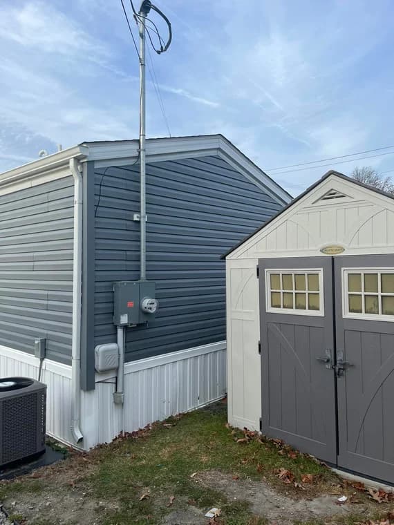 Exterior side of a house painted in dark gray siding with a white lower section, next to a beige storage shed with double doors.
