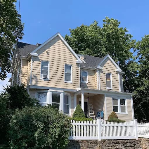 This is an image from Precision Painting NY, located in New York. The image shows a freshly painted two-story home exterior with tan siding, white trim, black roof shingles, and a white picket fence.