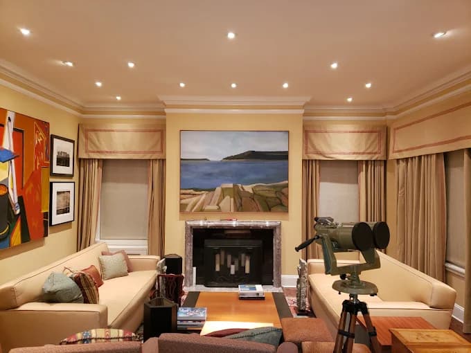 This is an image from Derheim Painting, located in Oklahoma. The photo shows a modern living room with beige walls, cream sofas, framed artwork, and recessed ceiling lights, creating a warm and inviting atmosphere.