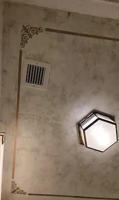 This is an image from Interior Finishing NYC, located in New York. The ceiling features a decorative faux-finish texture with ornate painted corner details, a recessed air vent, and a geometric ceiling light.