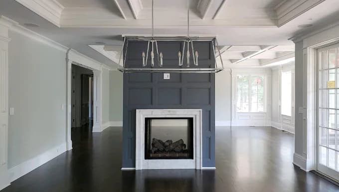 This is an image from Precision Painting Plus of Suffolk County, located in New York. The photo shows a modern living room with a dark gray accent wall surrounding a fireplace, coffered ceilings, and freshly painted white trim.