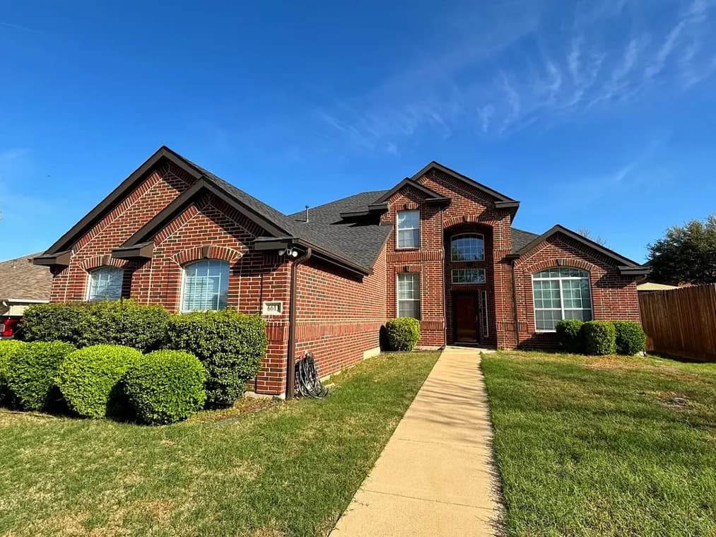 "This is an image from Roll City Painting, located in Texas. The exterior shows a red-brick home with dark trim, well-shaped shrubs, and a clear blue sky."