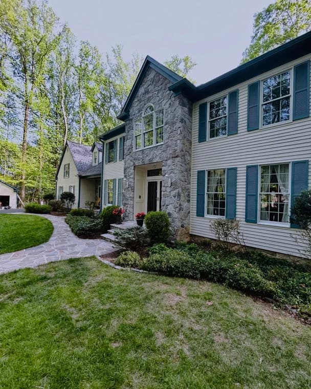 This is an image from C & V Painting Services LLC, located in Virginia. The exterior photo shows a large two-story home with stone and siding, blue shutters, and a landscaped walkway leading to the front entrance.
