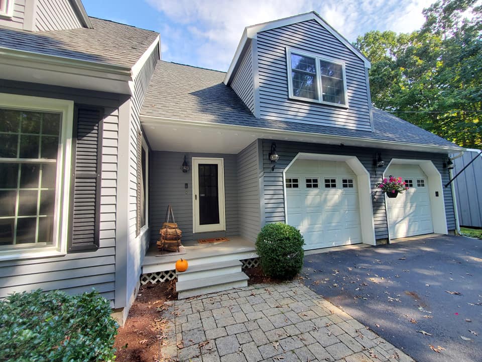 This is an image from WC Painting Inc, located in Massachusetts. The image shows a freshly painted gray home exterior with white trim, double garage doors, and black shutters, displaying a neat and clean finish.