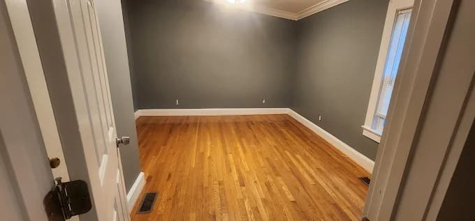 Interior painting project by Garner's Painting Service in Fayetteville, North Carolina. The image shows gray walls, white trim, wood flooring, a window, and ceiling light.