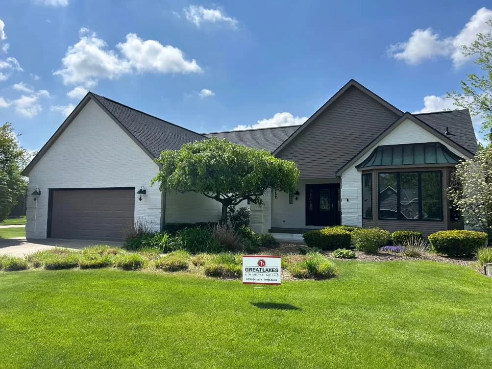 This is an image from Great Lakes 1-800 Painting LLC company, located in Davison, MI The image is a beautiful modern home with freshly painted white brick exterior, dark brown roof, and trim accents, completed by Great Lakes Painting — sign displayed on green lawn.