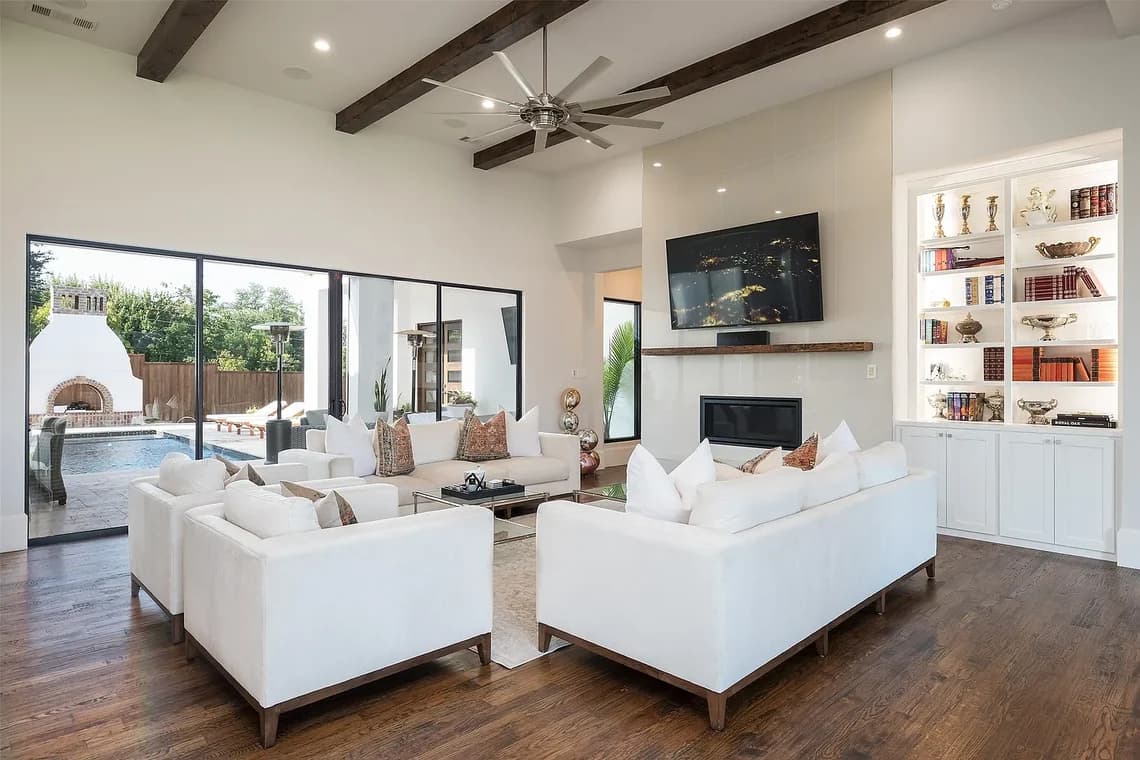 "This is an image from Highland Painting Co., located in Texas. The interior scene shows a spacious living room with white furniture, exposed ceiling beams, hardwood floors, and large sliding glass doors overlooking an outdoor patio and pool."