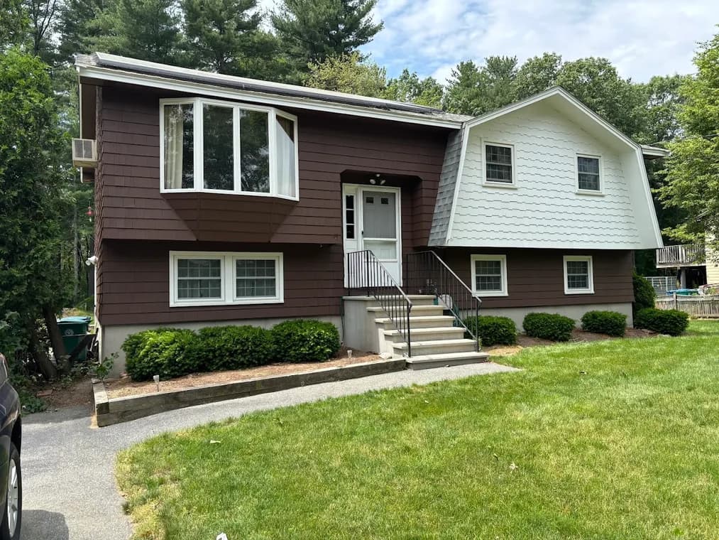 This is an image from Ortolani & Sons Painting Company, located in Massachusetts. The image shows a split-level home exterior with freshly painted brown and light gray siding, white trim, and a clean, updated finish surrounded by green landscaping.