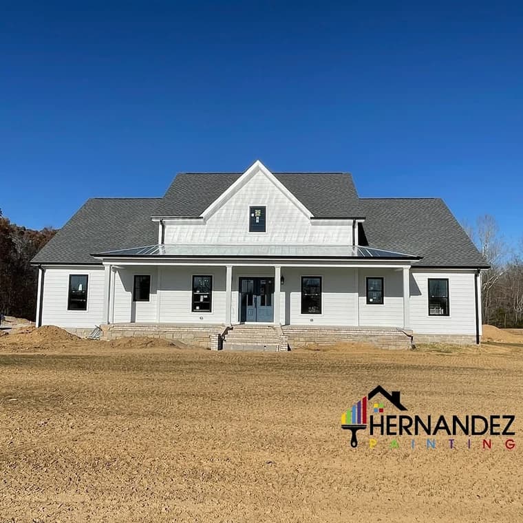 This is an image from Hernandez Painting, located in Tennessee. The photo shows a newly constructed white farmhouse-style home with black trim and a dark gray roof under a clear blue sky.