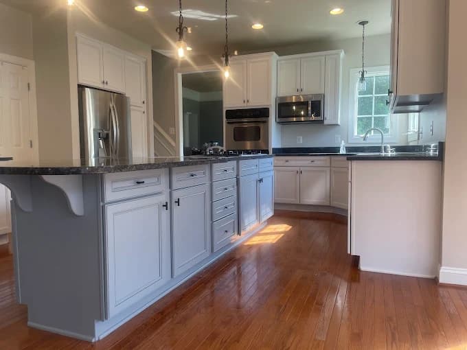 This is an image from C&R Pro Painting, located in Virginia. The image shows a spacious kitchen with white cabinetry, gray island cabinets, dark stone countertops, pendant lighting, and polished hardwood floors.