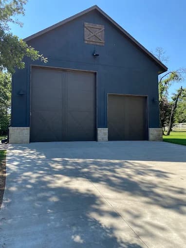 This is an image from Cardinal Painting, located in Oklahoma. The photo shows a large barn-style garage freshly painted in a deep charcoal color with light stone accents and a wide concrete driveway.