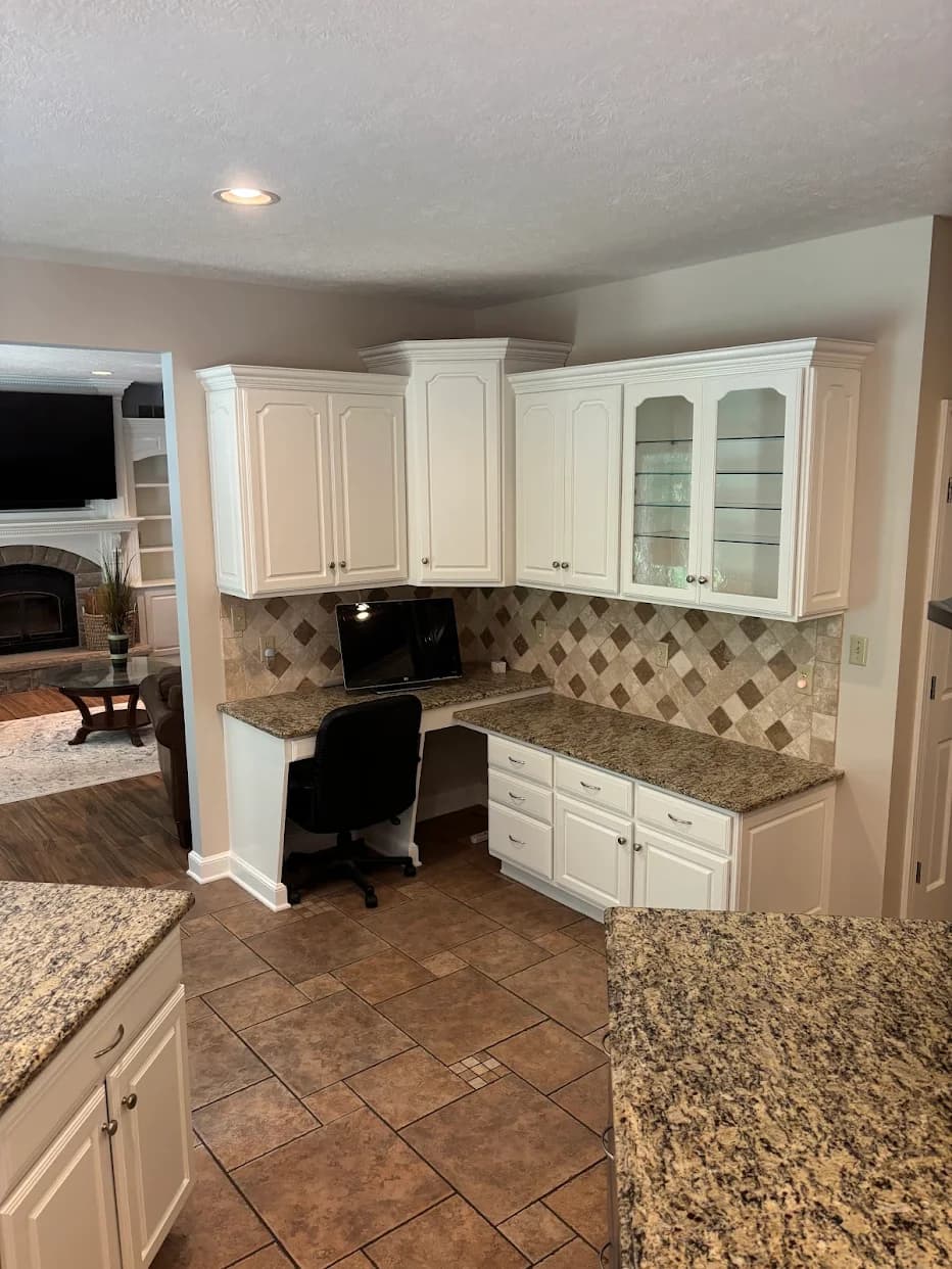 This is an image from Resurrection Painting LLC, located in Ohio. The image shows a freshly painted kitchen with white cabinets, decorative crown molding, granite countertops, tiled backsplash, and a built-in desk area with clean, neutral interior finishes.