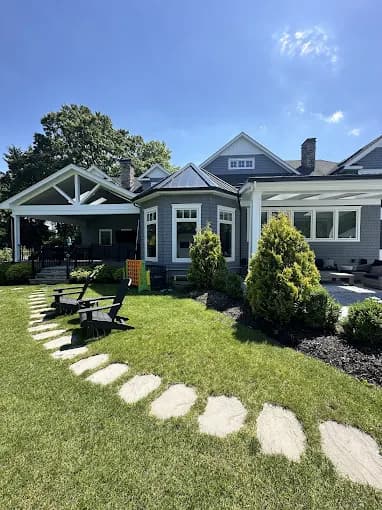 This is an image from Teall Painting, located in Michigan. The image shows a completed exterior painting project on a large residential home with gray siding, white trim, and a landscaped backyard patio area.