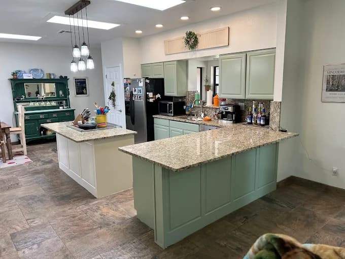 This is an image from Innovative Coatings LLC, located in New Mexico. The photo shows a remodeled kitchen with mint green lower cabinets, granite countertops, pendant lighting, and modern tile flooring extending into a bright dining area.
