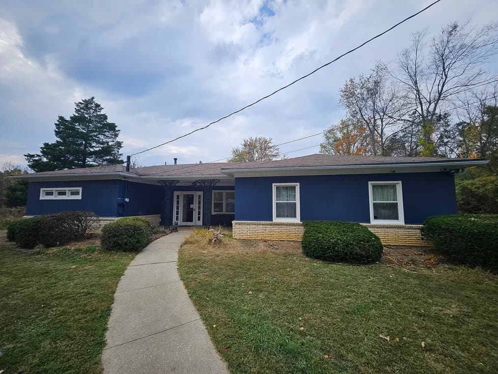 This is an image from Abell Painting LLC, located in Kentucky. The photo shows a single-story house with a fresh navy-blue exterior paint job and light brick trim, surrounded by trimmed bushes and trees under a cloudy sky.