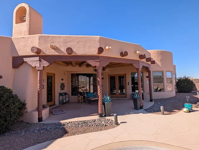 This is an image from Knight's Painting, located in New Mexico. The photo shows a Southwestern-style home exterior with light tan stucco walls, brown wooden beams, and a shaded patio area with desert landscaping.