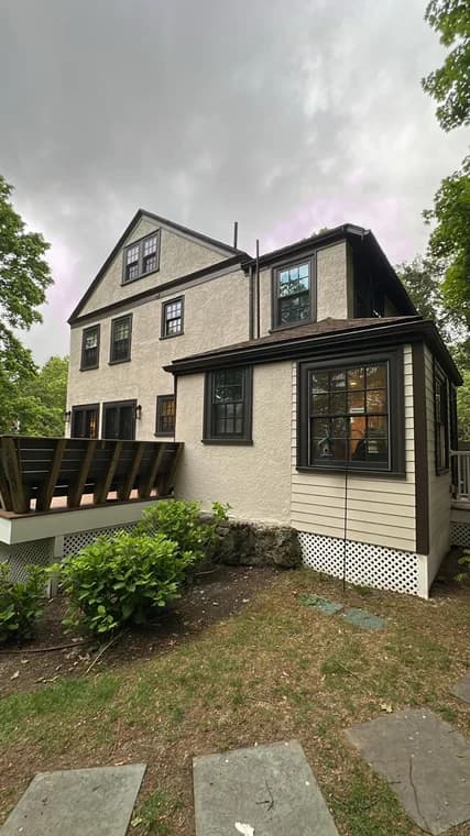 A large multi-story home with cream and brown trim freshly painted by Titanium Painting & Renovations. The property includes black window frames and an attached deck surrounded by greenery.