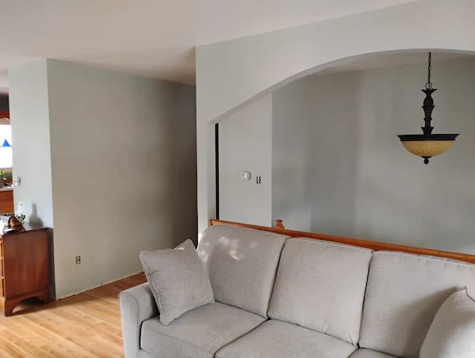 This is an image from Mendez Drywall & Painting LLC, located in Maryland. The image shows a living room with freshly painted light gray walls, an arched doorway, hardwood floors, and a neutral sectional sofa.