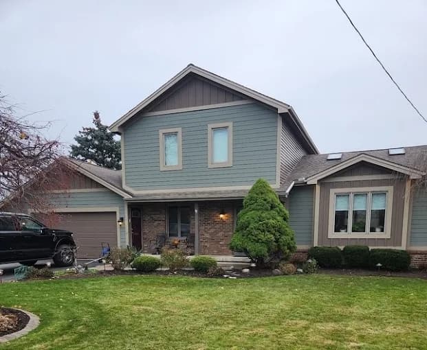 This is an image from R&R Painting & Concrete Coatings, located in New York. The exterior home photo shows newly painted gray siding with tan trim and brickwork on a two-story suburban house."