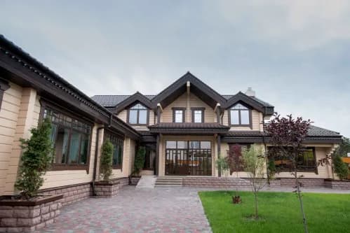 This is an image from The Woodlands Paint Doctor, located in Texas. The photo shows a large modern two-story home with light wood siding, dark roofing, and landscaped greenery along a paved walkway.