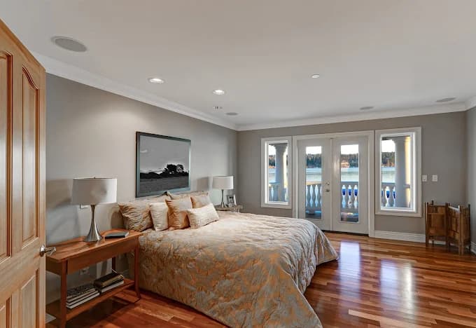 This is an image from Fresh Coat Painters of Oklahoma City, located in Oklahoma. The photo shows a spacious bedroom with freshly painted light gray walls, hardwood floors, a large bed with gold bedding, and French doors opening to a balcony view.