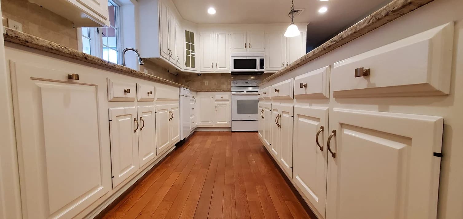 This is an image from WC Painting Inc, located in Massachusetts. The interior kitchen features newly painted white cabinets with brass handles, granite countertops, and polished wood flooring for a bright modern look.