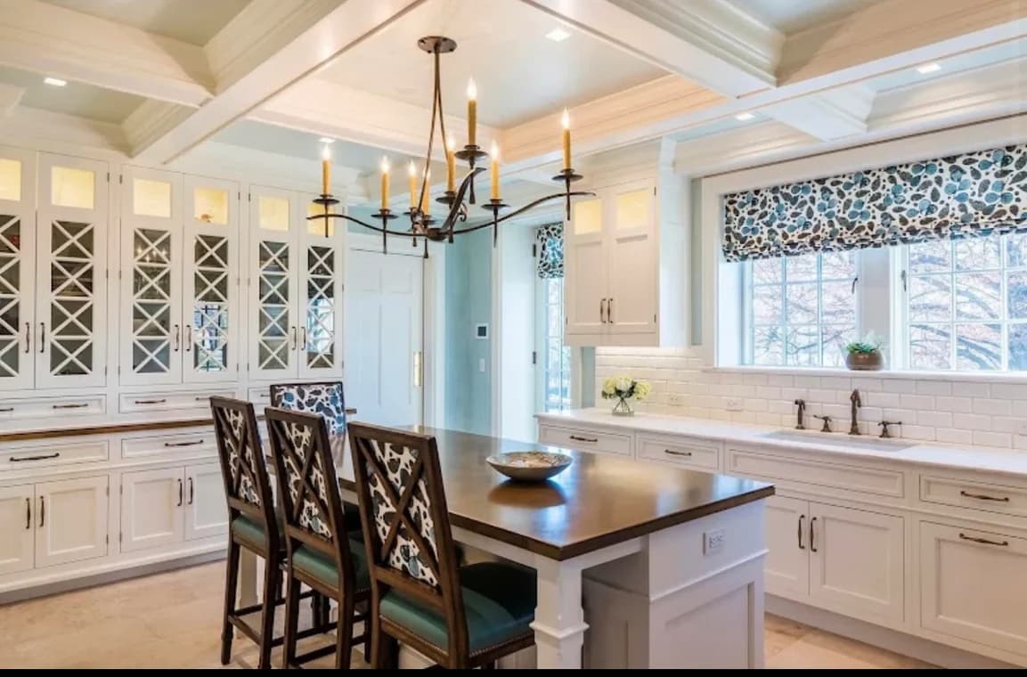 "This is an image from Prestige Pro Painters, located in Texas. The image displays a bright kitchen with white cabinetry, a large island, patterned window treatments, and a chandelier hanging over the workspace."